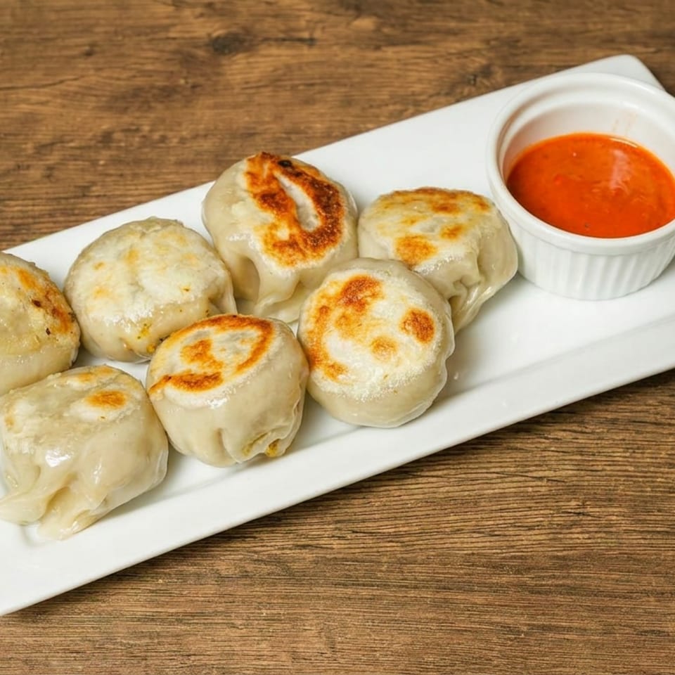 Seven pan-fried dumplings arranged on a white rectangular plate with a bowl of red dipping sauce.